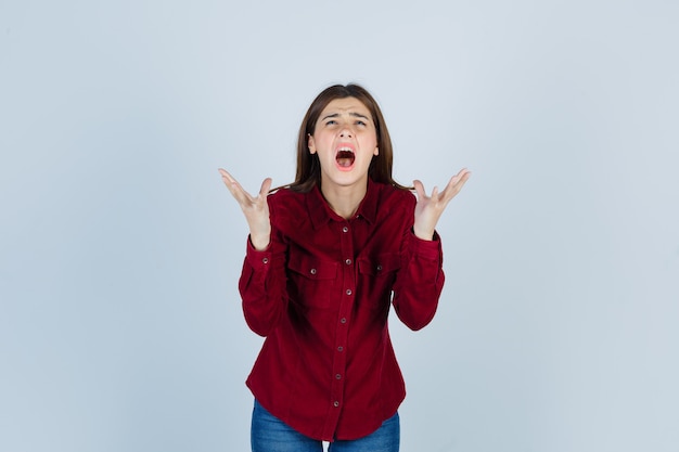 Free photo portrait of girl raising hands while screaming in burgundy shirt and looking wistful