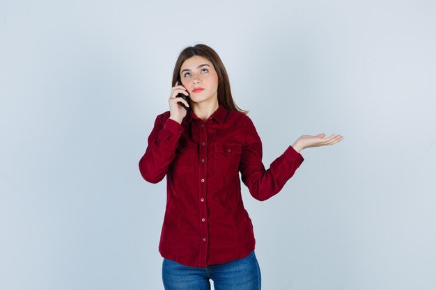 Portrait of girl showing helpless gesture while talking on smartphone in burgundy shirt and looking pensive