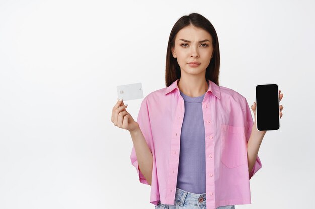 クレジットカードが眉をひそめ、白い背景の上に立って疑わしいように見える携帯電話の画面を示す女の子の肖像画
