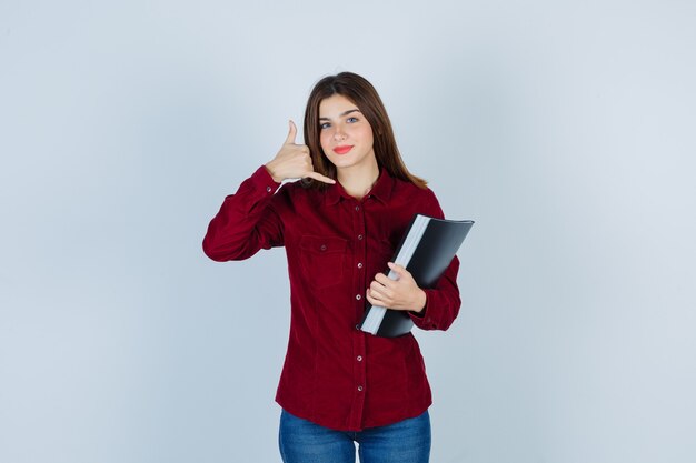 Portrait of girl showing phone gesture, holding folder in burgundy shirt and looking sincere