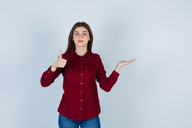 Portrait of girl showing thumb up, spreading palm aside in burgundy shirt and looking confident