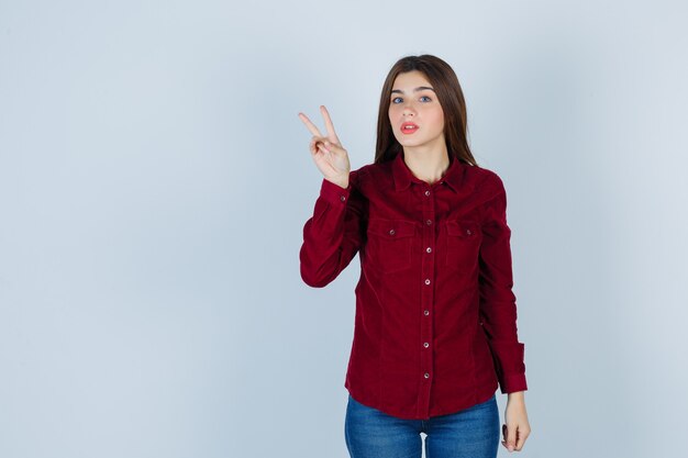 Portrait of girl showing victory gesture in burgundy blouse and looking puzzled