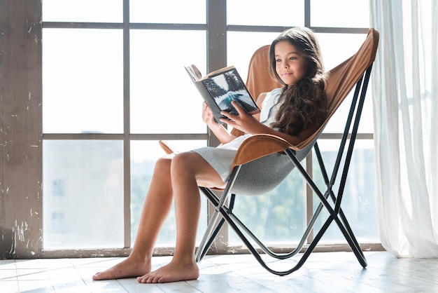 Free photo portrait of a girl sitting on chair near the window reading book