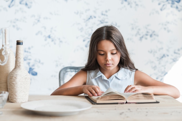 Free photo portrait of a girl sitting in front of wallpaper reading book