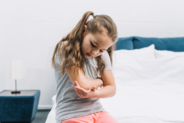 Portrait of a girl sitting on white bed looking at her injured elbow