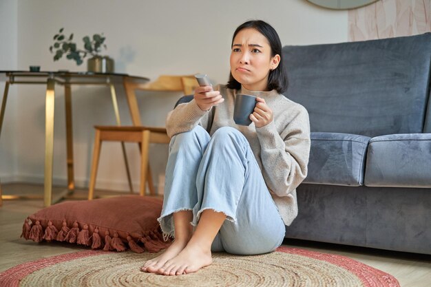 Portrait of girl watching television at home sits on floor near sofa holds remote and changes channe