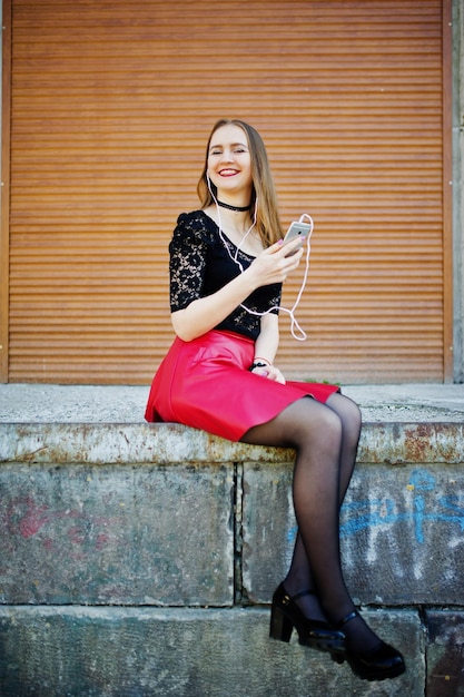 Free photo portrait of girl with black choker on her neck red leather skirt and mobile phone at hand with headphones against orange shutter