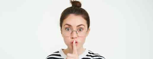 Free photo portrait of girl with glasses looks surprised hughing press finger to lips shush shh gesture asks to