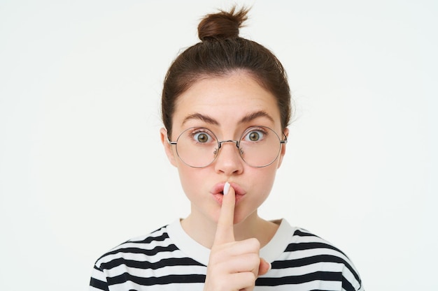 Free photo portrait of girl with glasses looks surprised hughing press finger to lips shush shh gesture asks to