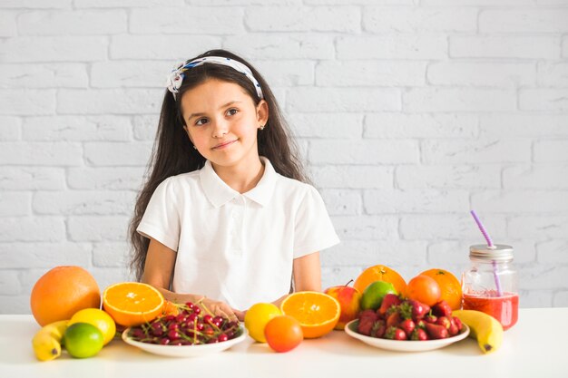Portrait of a girl with many fruits on white desk against wall