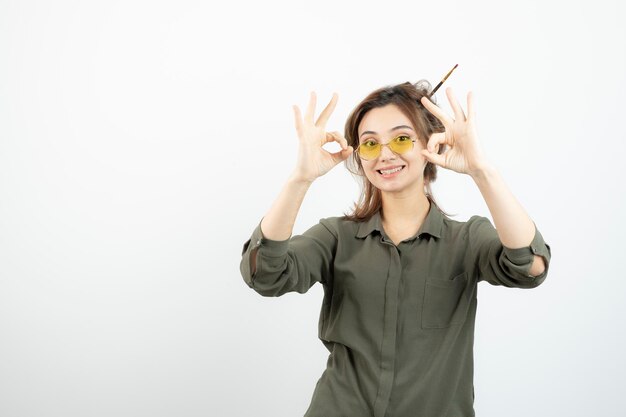 Portrait of girl with messy bun in glasses standing and giving ok sign. High quality photo