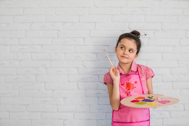 Portrait of a girl with paintbrush and palette standing against white wall