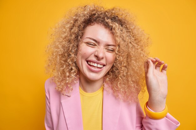 巻き毛のふさふさした髪の笑顔で嬉しいヨーロッパの女性の肖像画は、本物の感情を喜んで表現します黄色の壁に隔離されたエレガントな服に身を包んだ喜びから目を閉じてとても嬉しいです