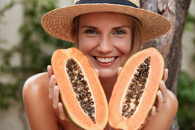 portrait of glad lovely woman poses naked, wears summer hat, holds organic exotic papaya