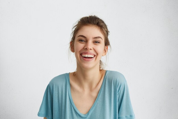 Портрет рад женщины, искренне улыбаясь. Европейская женщина с широкой улыбкой в синей повседневной блузке позирует у белой стены