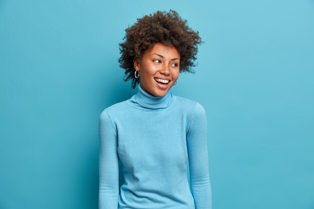 기쁜 젊은 아프리카 계 미국인 여성의 초상화는 넓게 웃고, 즐거운 표정을 짓고, 캐주얼 한 파란색 터틀넥을 입고, 머리를 옆으로 돌리고, 재미있는 장면을 발견합니다. 흑백 촬영. 행복 한 감정 개념