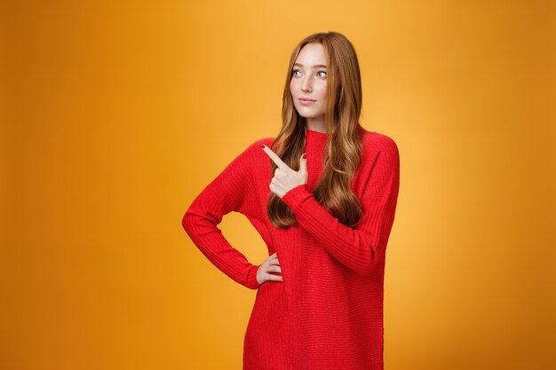 魅力的でスタイリッシュな赤毛の女性起業家の肖像画は、オレンジ色の壁を越えてビジネスを管理し、思慮深く見つめ、仕事に集中しているように左上隅を指している新しいプロジェクトを設計および主導しています