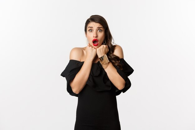 Portrait of glamour woman looking scared and shocked at camera, staring at something with fear, standing in black dress against white background.