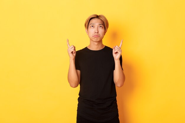 Portrait of gloomy asian blond man, wearing black t-shirt, sulking disappointed and pointing fingers up, yellow wall.