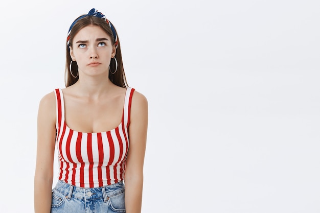 Portrait of gloomy upset attractive european woman in headband and striped top pursing lips and looking up envy or unhappy over grey wall