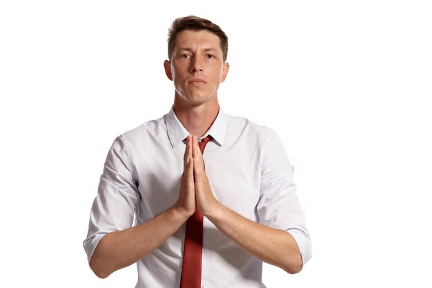 Portrait of a good brunet guy with brown eyes, wearing in a white shirt and a red tie. He is praying while posing in a studio isolated over a white background. Concept of gesticulation and sincere emo