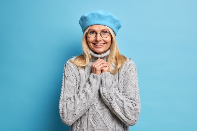 Portrait of good looking adult European woman smiles pleasantly, keeps hands together, wears optical glasses, french beret and knitted sweater