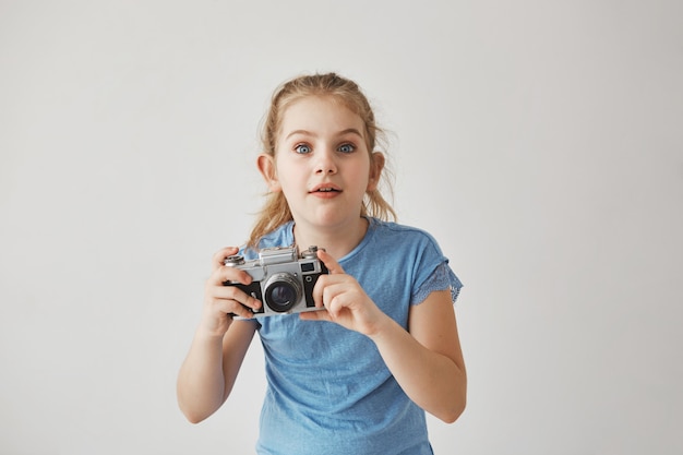 Free photo portrait of good-looking blond girl in blue t-shirt holding camera in hands with concentrated expression, going to take a picture of cute cat on street.