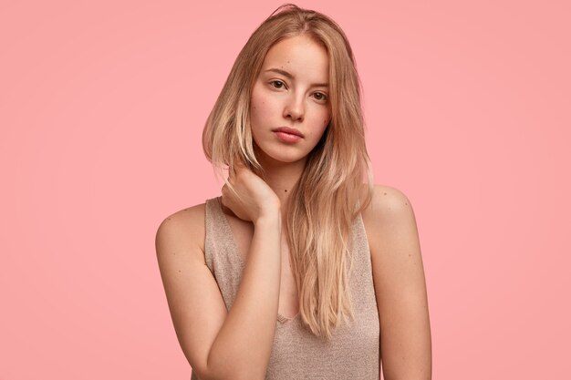 Портрет красивой блондинки касается шеи руками, выглядит загадочно и серьезно, у нее чистая кожа, она собирается гулять на свежем воздухе, модели у розовой стены
