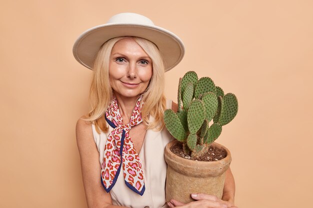 Portrait of good looking blonde woman with minimal makeup well cared comlexion holds pot of cactus wears hat dress tied kerchief isolated over beige wall