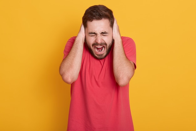 Portrait of good looking caucasian guy in red t shirt, male yelling with closed eyes and covering ears palms, not want to hear noise