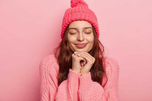 Ritratto di bella donna affascinante tiene entrambe le mani sotto il mento, gli occhi chiusi, essendo immerso nei pensieri, modelli sopra il muro roseo dello studio, vestito con maglione e cappello