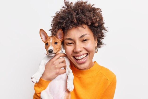 Portrait of good looking cheerful African American woman enjoys company of small pedigree dog wears orange jumper spends free time with favorite pet isolated over white wall. Animal owner