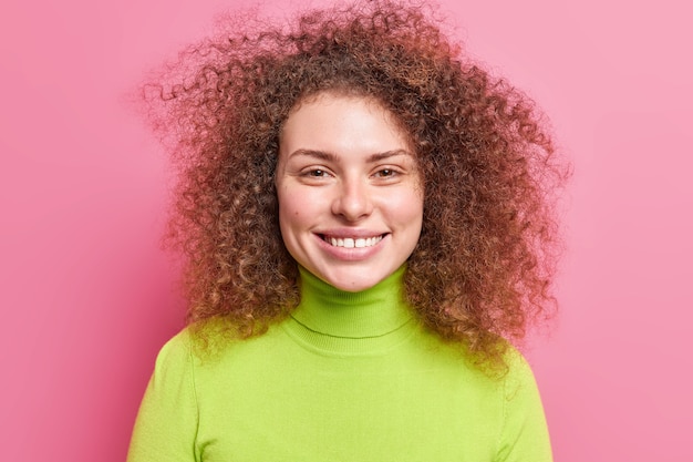 Portrait of good looking cheerful European woman with curly bushy hair smiles broadly dressed in green turtleneck isolated over pink wall. Carefree smiling European female enjoys free time