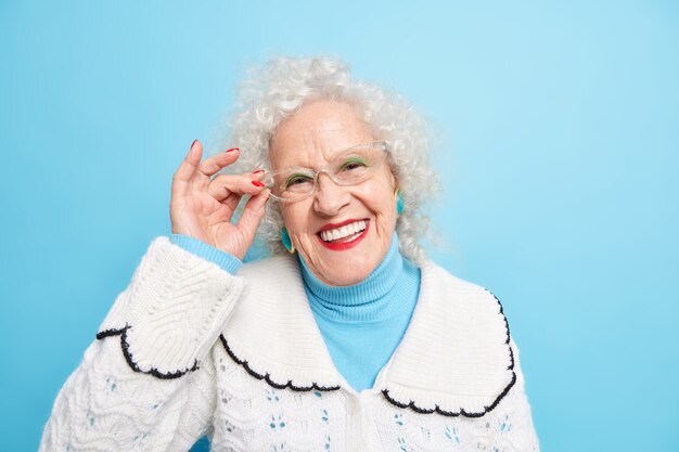 좋은 찾고 쾌활한 회색 머리 할머니의 초상화는 이빨로 안경의 가장자리에 손을 유지합니다 흰색 점퍼를 입고 잘 돌보는 안색 주름진 죄가 있습니다