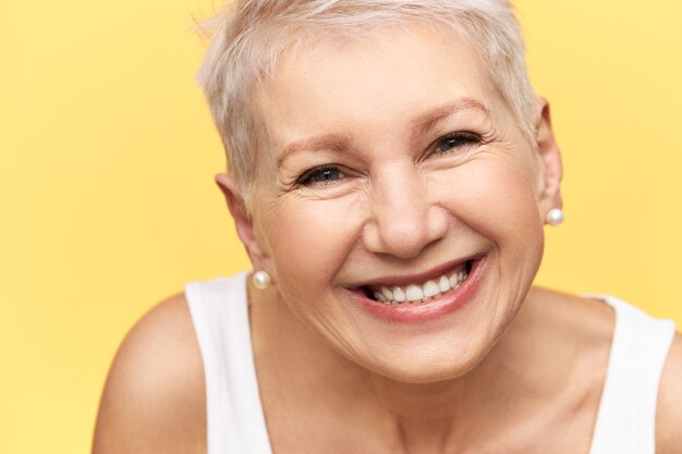 Portrait of good looking cheerful middle aged European female with stylish hairdo wearing white tank, expressing positive emotions, smiling broadly, showing straight teeth, happy to receive good news