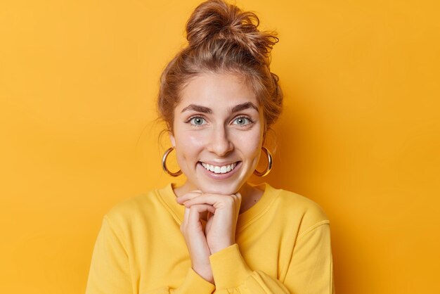 Portrait of good looking cheerful woman keeps hands under chin smiles toothily stands happy indoors wears earrings and casual sweatshirt isolated over yellow background Positive emotions concept