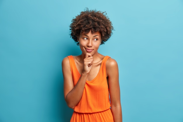 Portrait of good looking curly haired woman holds chin and looks with dreamy thoughtful expression aside