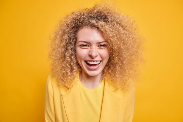 Portrait of good looking curly haired young woman smiles broadly expresses positive emotions glad to hear recent news 