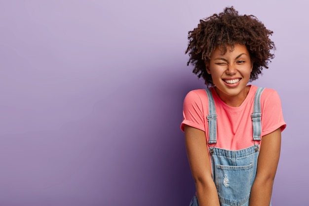 Portrait of good looking dark skinned woman with curly hairdo, blinks eye, has fun, smiles pleasantly, dressed in stylish clothes, expresses happy emotions, isolated over purple background, copy space