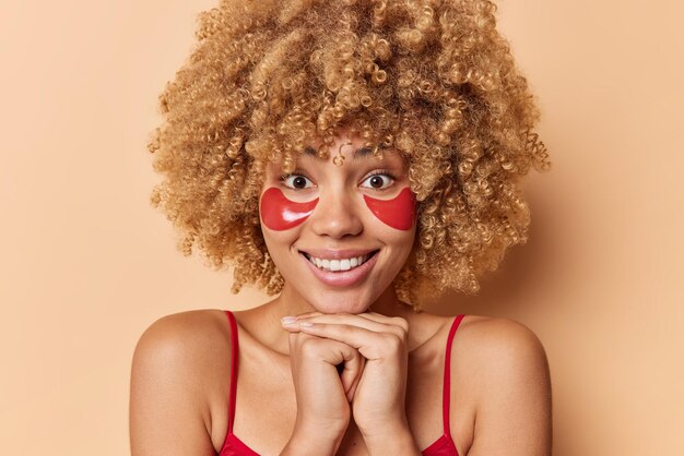 巻き毛のふさふさした髪の笑顔を持つ格好良いヨーロッパの女性の肖像画は、あごの下に手を心地よく保ち、茶色の背景の上にさりげなく隔離された服を着た目の下に赤いヒドロゲルパッチを適用します