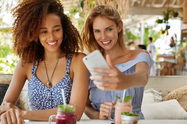 Портрет симпатичных женщин делает селфи на смартфон, сидит вместе в летнем кафе, проводит летние каникулы за границей, довольные хорошо отдохнуть. Люди, общение и отношения