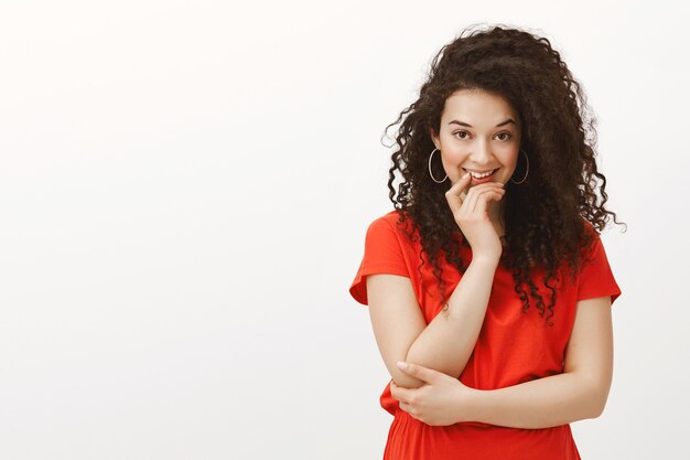 Portrait of good-looking flirty femenine woman with curly hair, smiling intriguingly and holding hand on ching