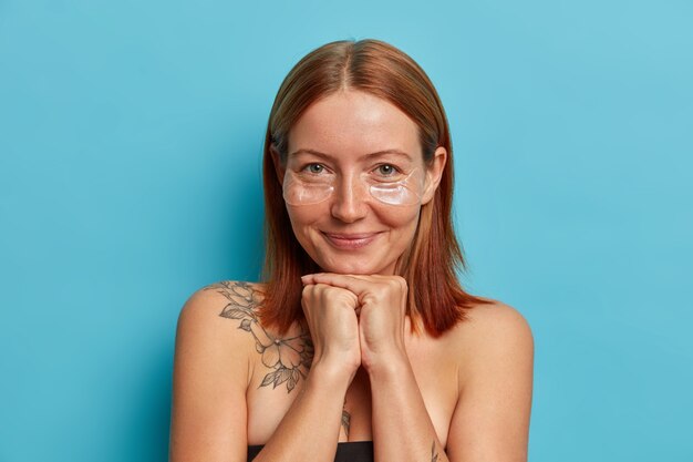 Portrait of good looking freckled redhead woman keeps hands under chin, applies transparent collagen patches under eyes, hydrates skin and removes wrinkles, poses with bare shoulders 