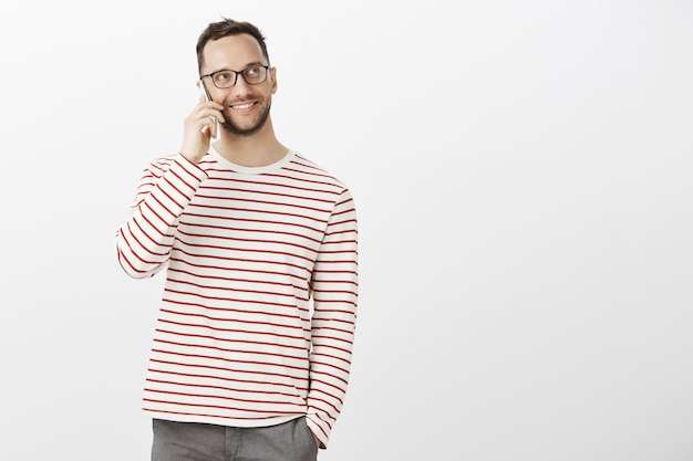 Portrait of good-looking friendly male in trendy glasses and casual outfit, looking up while calling friend via smartphone