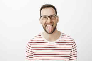Free photo portrait of good-looking funny adult coworker in black glasses, sticking out tongue and smiling joyfully, being carefree and careless about rules or duties