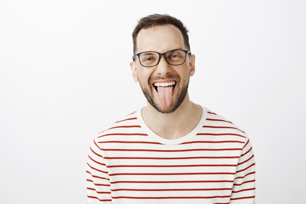 Portrait of good-looking funny adult coworker in black glasses, sticking out tongue and smiling joyfully, being carefree and careless about rules or duties