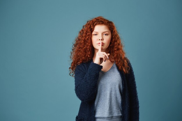 Portrait of good-looking ginger student woman with wavy hair holding index finger in front of lips showing friend not to tell anyone story she just said.