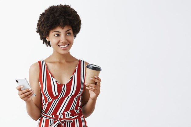 Portrait of good-looking girly african american female with curly hairstyle holding cup of coffee and smartphone gazing right with cute smile