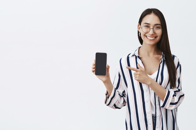 Портрет красивой довольной модной женщины в очках и блузке, показывающей экран на телефоне и указывающей на устройство указательным пальцем, широко улыбаясь