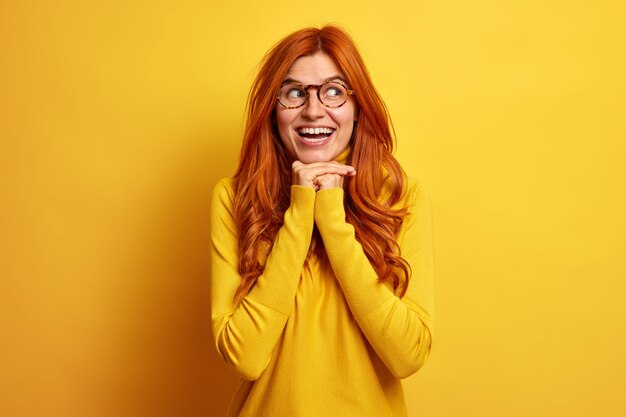 Portrait of good looking redhead European woman smiles broadly keeps hands under chin looks aside gladfully wears casual jumper expresses happiness.